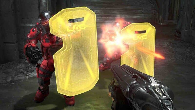 Một cảnh bắn súng trong DOOM Eternal Ảnh gameplay game DOOM Eternal, góc nhìn thứ nhất, nhân vật chính đang bắn một con quỷ màu đỏ với một khẩu súng máy. Con quỷ đang cầm một tấm khiên năng lượng màu vàng, cho thấy cơ chế chiến đấu phức tạp của game.
