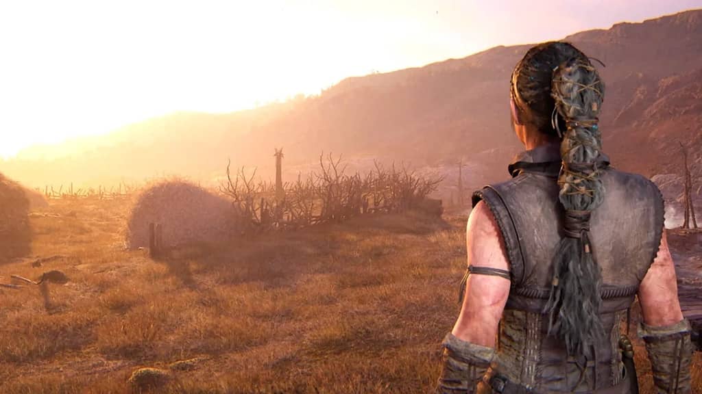 Bình minh u ám trong Senua's Saga: Hellblade II Enhanced. Senua, với mái tóc bện đặc trưng và vẻ mặt kiên định, đứng quay lưng về phía người xem trên một vùng đất cằn cỗi, phía trước là một hình bóng khổng lồ và bí ẩn ẩn hiện trong màn sương mù và ánh sáng vàng vọt của bình minh. Khung cảnh gợi lên cảm giác về một hành trình đầy gian truân và những thử thách đáng sợ đang chờ đợi.