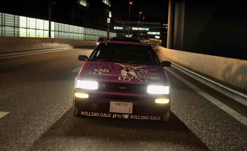 Chiêm ngưỡng chiếc xe drift ấn tượng trong Tokyo Xtreme Racer. Tokyo Xtreme Racer, chiếc xe drift màu hồng có decal Rolling Gals đang chạy trên đường cao tốc.